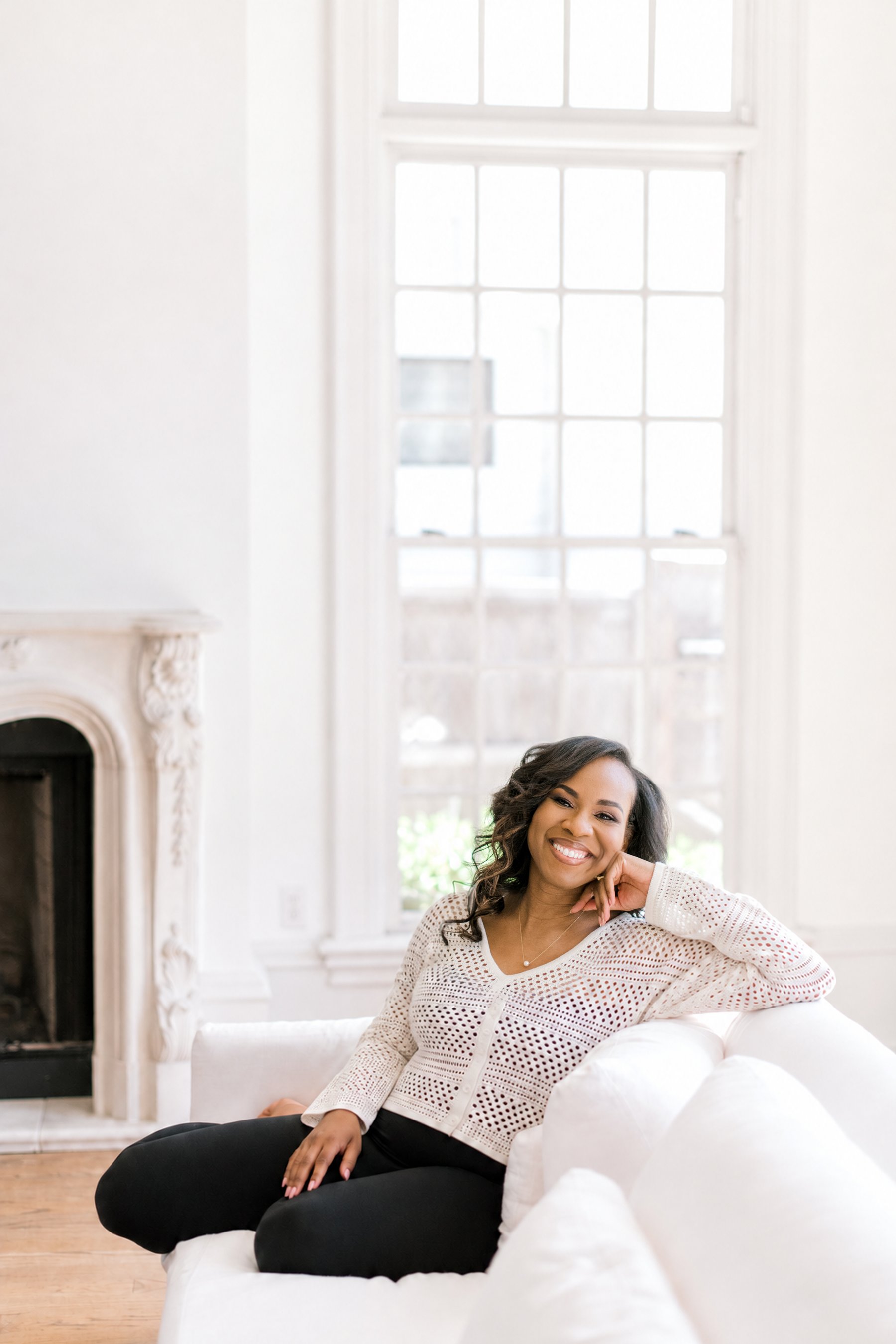 Amie Johnson smiling, seated on a white sofa in front of a tall paned window